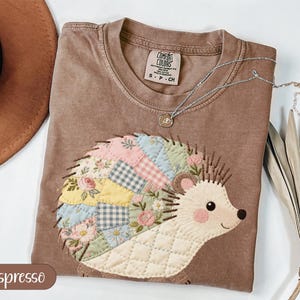 May include: Espresso-colored t-shirt with a hedgehog applique. The hedgehog has a patchwork design with floral and geometric patterns in pastel colors. The shirt has a crew neck and a small tag. A silver necklace with a pendant is on the shirt.