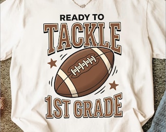 Puede incluir: Una camiseta de manga corta color blanco hueso con un gráfico de un balón de fútbol americano marrón en el centro. El texto READY TO TACKLE 1ST GRADE aparece en letras mayúsculas marrones. Dos pequeñas estrellas marrones flanquean el balón.