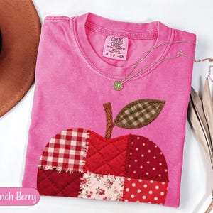 May include: A bright pink short-sleeve t-shirt featuring a large apple-shaped patch made of various red and white fabric squares with gingham, polka dot, and floral patterns. The apple has a brown stem and a green gingham leaf with visible stitching.