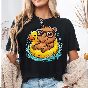 Peut inclure: Un t-shirt noir avec une illustration graphique d'un capybara brun portant des lunettes à monture noire, assis dans une bouée de piscine jaune vif en forme de canard, entouré d'éclaboussures d'eau bleues et de petites bulles blanches.