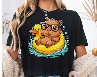 Puede incluir: Una camiseta negra con una ilustración gráfica de un capibara marrón con gafas de montura negra, sentado dentro de un flotador amarillo brillante con forma de pato, rodeado de salpicaduras de agua azules y pequeñas burbujas blancas.