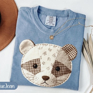 Könnte beinhalten: Ein blaues Jeans-T-Shirt mit einem Patchwork-Waschbärgesicht-Design. Das Waschbärgesicht besteht aus verschiedenen Stoffen, darunter Blumen- und Karomuster. Eine goldene Halskette mit einem kleinen Anhänger liegt auf dem Shirt. Das Shirt hat ein Comfort Colors-Etikett.