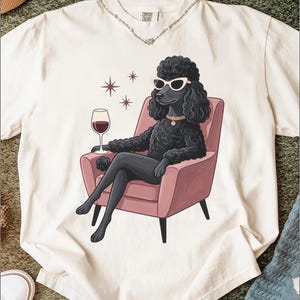 Può includere: Una t-shirt a maniche corte color bianco sporco con la grafica di un barboncino nero che indossa occhiali da sole bianchi a gatto e un collare. Il barboncino è seduto su una poltrona rosa, tiene un calice di vino rosso e tre stelline.