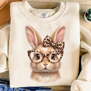 Puede incluir: Sudadera de color crema con una ilustración detallada de un conejito con gafas y un lazo. El conejito tiene pelaje marrón, un lazo con estampado de leopardo y gafas con montura de leopardo. La sudadera es de estilo casual.
