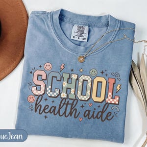 Puede incluir: Camiseta azul claro con la frase "SCHOOL health aide" en un diseño retro colorido. El diseño incluye caras sonrientes, flores y rayos. Un collar dorado está drapeado sobre la camiseta.