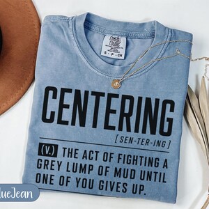 May include: A light blue t-shirt with the word "CENTERING" in large black letters. Below the word is a definition that reads "THE ACT OF FIGHTING A GREY LUMP OF MUD UNTIL ONE OF YOU GIVES UP."