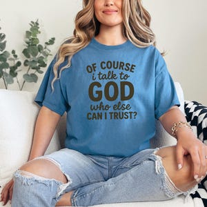 May include: A blue t-shirt with the text "OF COURSE i talk to GOD who else CAN I TRUST?" in brown lettering. The shirt is worn with ripped blue jeans. The person is sitting on a white couch.