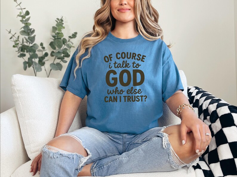 May include: A blue t-shirt with the text "OF COURSE i talk to GOD who else CAN I TRUST?" in brown lettering. The shirt is worn with ripped blue jeans. The person is sitting on a white couch.