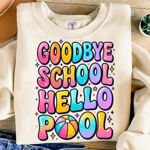 May include: A cream-colored crewneck sweatshirt featuring a colorful graphic print with the text "GOODBYE SCHOOL HELLO POOL" in a bubble-style font. The design includes a beach ball graphic and small star accents in shades of pink, yellow, and blue.