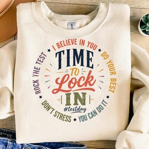Puede incluir: Sudadera de color crema con un diseño circular que dice "I believe in you, Time to Lock In, Do Your Best, You Can Do It." El texto es en varios colores, incluyendo azul marino, rojo y naranja. También incluye el hashtag #testday.