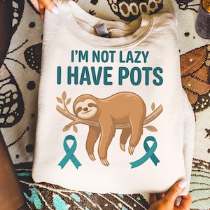 Puede incluir: Sudadera de color crema con el texto "I'M NOT LAZY I HAVE POTS" en color turquesa. Un perezoso de dibujos animados descansa sobre una rama debajo del texto, con cintas de concienciación turquesas a cada lado. La sudadera está doblada.