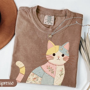 May include: Espresso-colored t-shirt featuring a patchwork cat design with pink, yellow, and blue accents. The cat has a whimsical, stitched appearance. A silver necklace with a small pendant rests on the shirt. The shirt is labeled "Comfort Colors".
