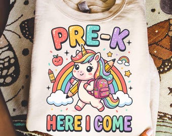 Puede incluir: Una sudadera beige de cuello redondo con un gráfico colorido de un unicornio con mochila, un arcoíris, una manzana y un lápiz. El texto dice PRE-K en la parte superior y HERE I COME en la inferior con letras redondeadas y multicolores.