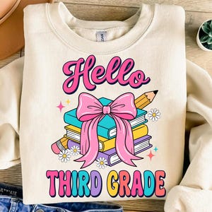 Peut inclure: Un sweat-shirt à col rond couleur crème avec une impression graphique représentant une pile de trois livres bleu sarcelle, jaunes et violets, noués par un grand nœud rose. Un crayon et des petites fleurs blanches entourent les livres.