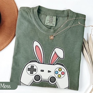 Könnte beinhalten: Ein moosgrünes T-Shirt mit einem weißen Controller-Design mit Hasenohren. Der Controller hat bunte Knöpfe und ein kreuzförmiges Steuerkreuz. Eine goldene Halskette liegt über dem Shirt.