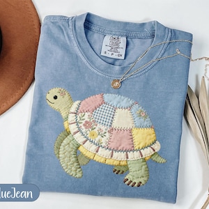 Peut inclure: Un t-shirt bleu clair Comfort Colors avec un motif de tortue en patchwork coloré. La carapace de la tortue présente un motif floral sur fond bleu. Un collier en or avec un pendentif est également visible.