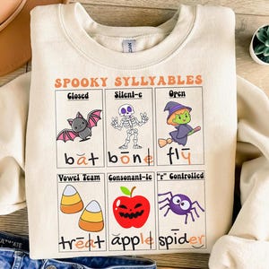 May include: Cream-colored sweatshirt with the text "SPOOKY SYLLYABLES" and illustrations of a bat, skeleton, witch, candy corn, apple, and spider. The sweatshirt has a round neckline and long sleeves.