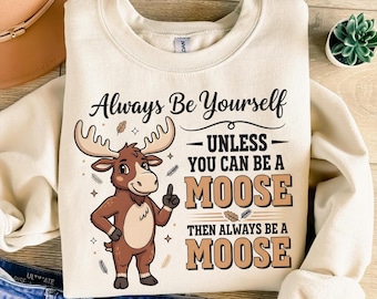 Puede incluir: Sudadera de color crema con el texto "Always Be Yourself - Unless You Can Be A Moose, Then Always Be A Moose." En la parte delantera se muestra una ilustración de un alce de dibujos animados, con colores marrón y beige.