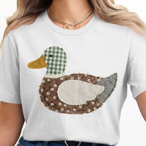 Könnte beinhalten: Weißes T-Shirt mit einem Enten-Applikation. Die Ente hat einen grün-weiß karierten Kopf, einen gelben Schnabel und einen Körper aus braunem Stoff mit weißen Blumenmustern. Der Flügel ist aus weißem Cord und der Schwanz ist grau.