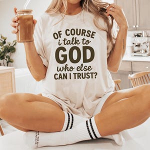 May include: Cream-colored t-shirt with the text "OF COURSE I talk to GOD who else CAN I TRUST?" in dark brown. The person is wearing white socks with black stripes and holding a glass of iced coffee.