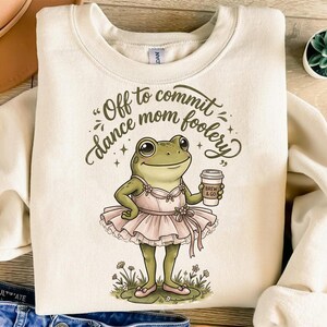 Könnte beinhalten: Cremefarbenes Sweatshirt mit einem skurrilen Frosch-Ballerina-Design. Der Frosch trägt einen rosa Tütü und hält eine Kaffeetasse mit der Aufschrift "Brew & Go". Der Text über dem Frosch lautet "Off to commit dance mom foolery."