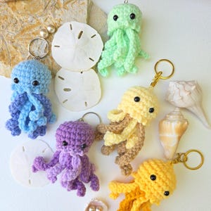 May include: A collection of handmade crochet jellyfish keychains in various colors, including blue, green, yellow, and purple. Each keychain features a metal ring and a detailed, textured design, with black bead eyes. The keychains are displayed with seashells and sand dollars.