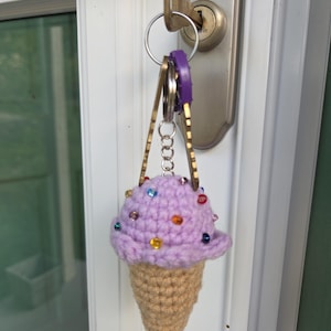 Op de afbeelding: Een gehaakte ijsje sleutelhanger. Het lavendelkleurige ijsje heeft kleurrijke kraal sprinkles, en de kegel is bruin. De sleutelhanger is bevestigd aan een set sleutels.