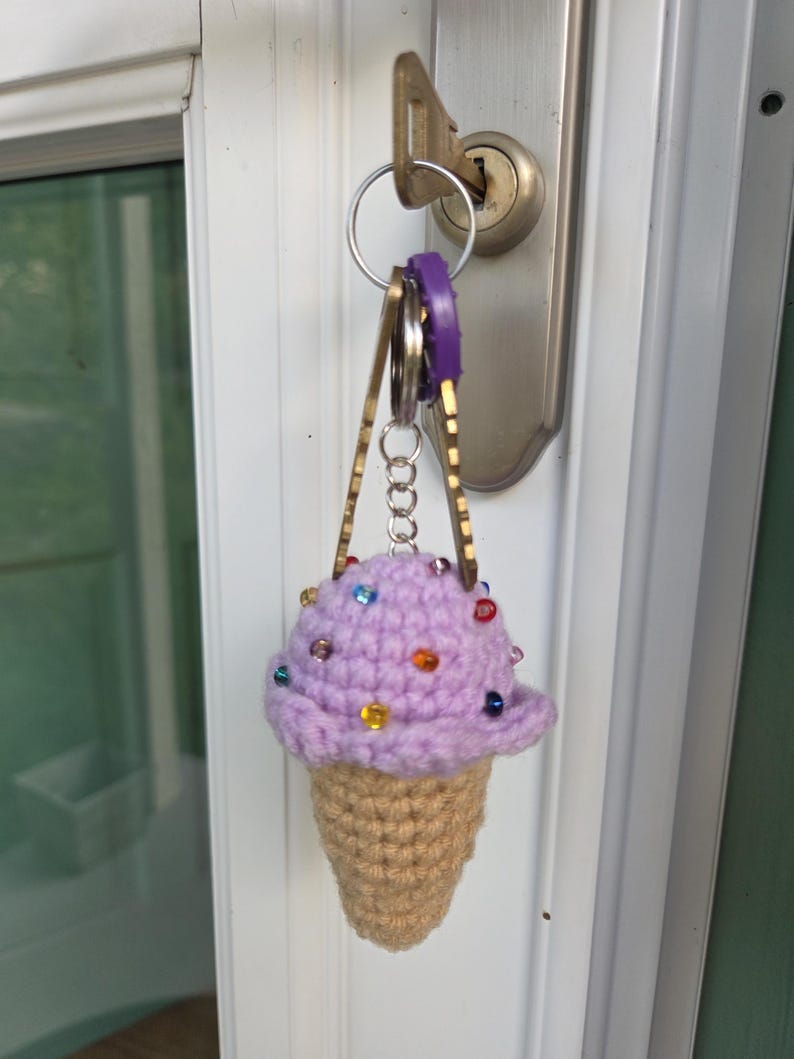 Op de afbeelding: Een gehaakte ijsje sleutelhanger. Het lavendelkleurige ijsje heeft kleurrijke kraal sprinkles, en de kegel is bruin. De sleutelhanger is bevestigd aan een set sleutels.