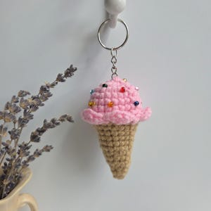 Op de afbeelding: Een gehaakte ijsje sleutelhanger. Het roze ijsje heeft kleurrijke kraal sprinkles en de kegel is lichtbruin. De sleutelhanger is bevestigd aan een zilveren metalen ring. De achtergrond is een effen witte muur.