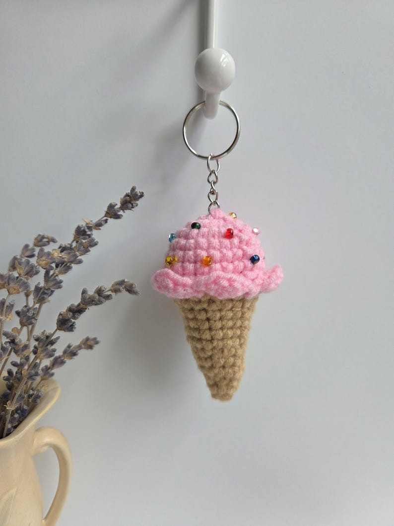 Op de afbeelding: Een gehaakte ijsje sleutelhanger. Het roze ijsje heeft kleurrijke kraal sprinkles en de kegel is lichtbruin. De sleutelhanger is bevestigd aan een zilveren metalen ring. De achtergrond is een effen witte muur.