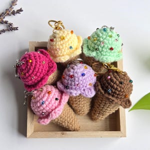 Op de afbeelding: Een verzameling van zes gehaakte ijsjessleutelhangers in een houten dienblad. De ijsbollen zijn in verschillende kleuren, waaronder roze, geel, groen, paars en bruin, en zijn versierd met kleurrijke kralen. De kegels zijn lichtbruin.
