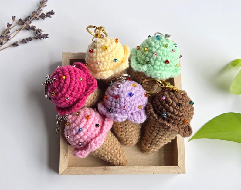 Op de afbeelding: Een verzameling van zes gehaakte ijsjessleutelhangers in een houten dienblad. De ijsbollen zijn in verschillende kleuren, waaronder roze, geel, groen, paars en bruin, en zijn versierd met kleurrijke kralen. De kegels zijn lichtbruin.
