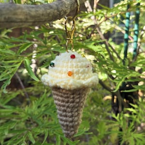 Op de afbeelding: Een handgemaakte gehaakte ijsje sleutelhanger. Het ijs is gebroken wit met kleurrijke kraal sprinkles, en de kegel is bruin. De sleutelhanger heeft een goudkleurige sluiting en ring, hangend aan een tak.