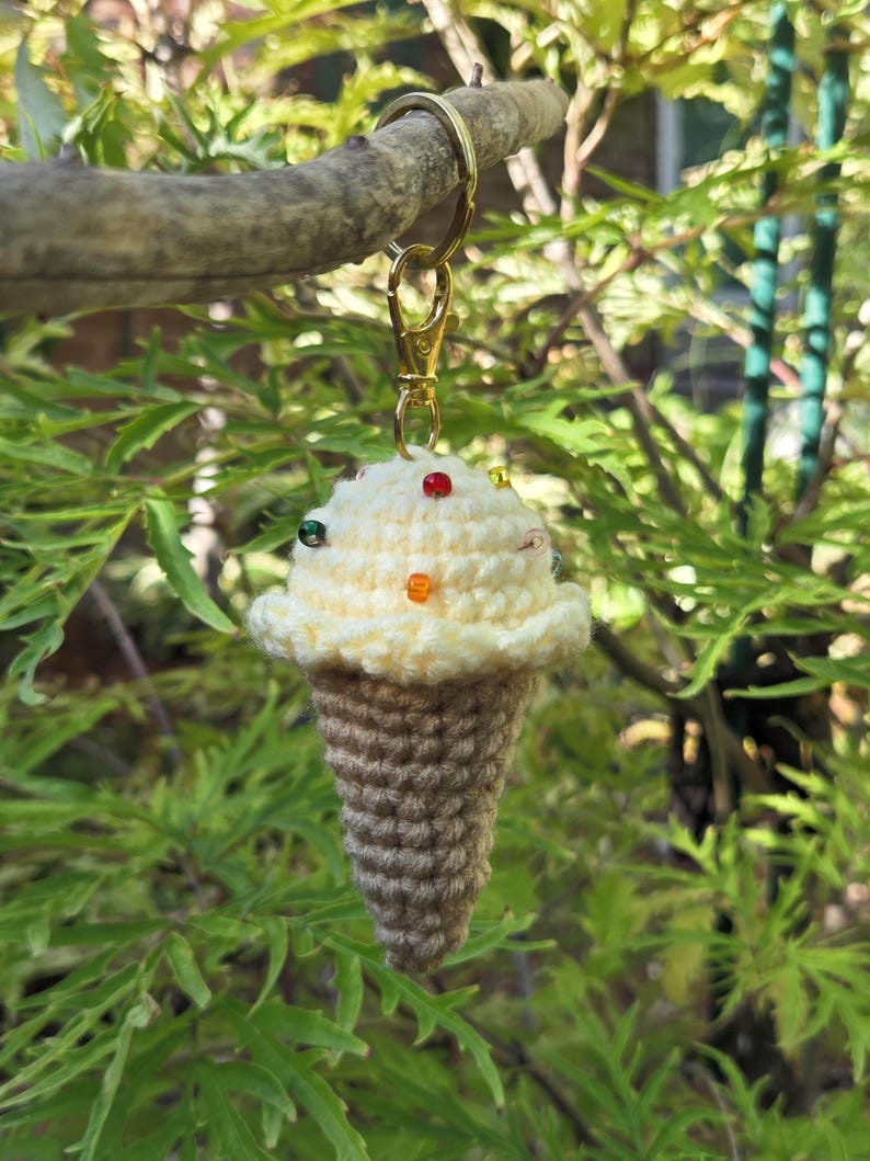 Op de afbeelding: Een handgemaakte gehaakte ijsje sleutelhanger. Het ijs is gebroken wit met kleurrijke kraal sprinkles, en de kegel is bruin. De sleutelhanger heeft een goudkleurige sluiting en ring, hangend aan een tak.