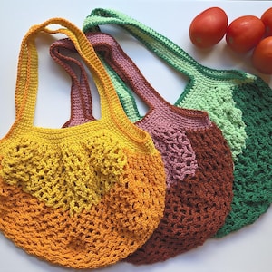 May include: Three handmade crochet market bags in shades of yellow, orange, brown, and green. The bags have open, net-like designs and sturdy handles. Fresh red tomatoes are visible in the background, suggesting the bags' use for carrying produce.