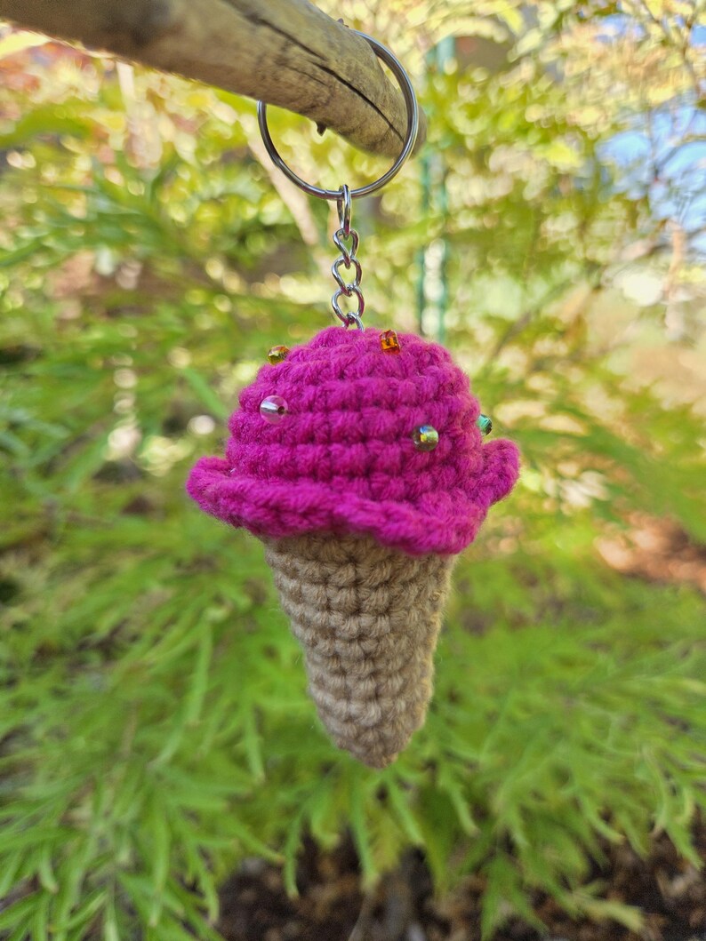 Op de afbeelding: Een handgemaakte gehaakte ijsje sleutelhanger. Het ijs is felroze, versierd met kleine, kleurrijke kralen. De kegel is lichtbruin van kleur. De sleutelhanger is bevestigd aan een zilveren metalen ring.