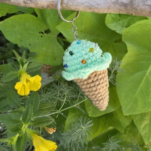 Op de afbeelding: Een handgemaakte gehaakte ijsje sleutelhanger. Het ijs is mintgroen met kleurrijke kraal sprinkles, en de wafel is bruin. De sleutelhanger is bevestigd aan een zilveren metalen ring. De achtergrond toont groen gebladerte en gele bloemen.