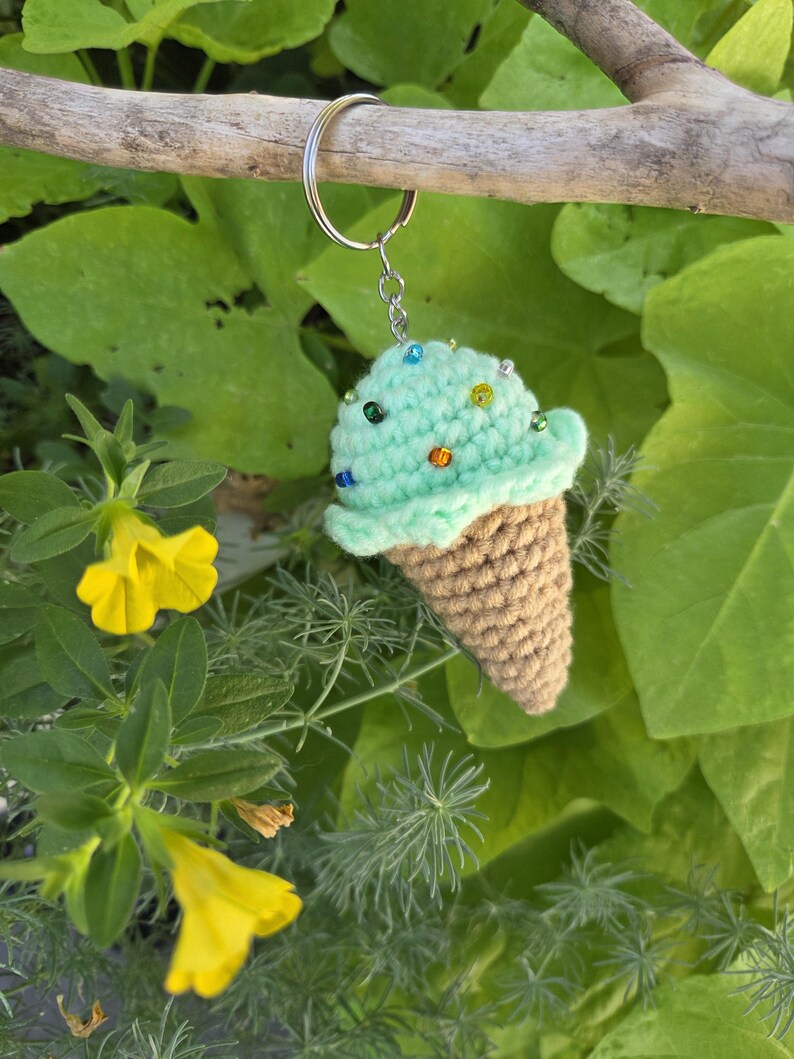 Op de afbeelding: Een handgemaakte gehaakte ijsje sleutelhanger. Het ijs is mintgroen met kleurrijke kraal sprinkles, en de wafel is bruin. De sleutelhanger is bevestigd aan een zilveren metalen ring. De achtergrond toont groen gebladerte en gele bloemen.