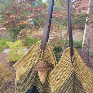 Op de afbeelding: Een handgemaakte gehaakte tote bag met een bruine leren riem. De tas heeft een kleurengradatie van olijfgroen naar marineblauw. Een kleine gehaakte ijsje-hanger hangt aan de riem.