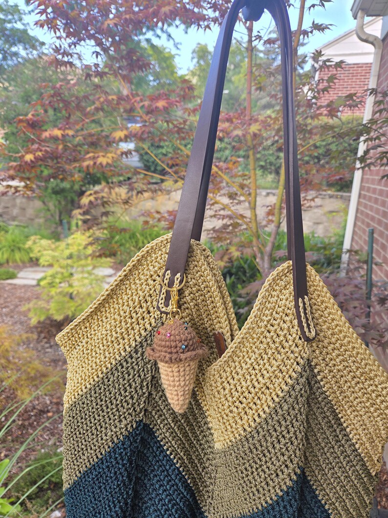Op de afbeelding: Een handgemaakte gehaakte tote bag met een bruine leren riem. De tas heeft een kleurengradatie van olijfgroen naar marineblauw. Een kleine gehaakte ijsje-hanger hangt aan de riem.