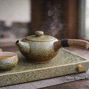 May include: A ceramic Kyusu teapot with a wooden handle and two matching cups, all on a speckled green tray. The teapot is steaming, suggesting hot tea. The text on the tray reads "240ml Kyusu Teapot."