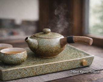 May include: A ceramic Kyusu teapot with a wooden handle and two matching cups, all on a speckled green tray. The teapot is steaming, suggesting hot tea. The text on the tray reads "240ml Kyusu Teapot."