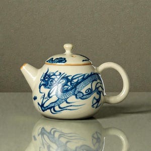 Op de afbeelding: Crèmekleurige keramische theepot met een blauw drakenontwerp. De theepot heeft een ronde body, een gebogen handvat en een deksel met een kleine knop. De draak is afgebeeld in een gedetailleerde, vloeiende stijl, die zich om het lichaam van de theepot wikkelt.