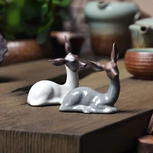 Peut inclure: Deux figurines de cerfs en céramique. L'une est blanche avec une tête et des oreilles marron, l'autre est grise avec une tête et des oreilles marron. Les deux sont en position de repos. Les figurines sont sur une surface en bois.