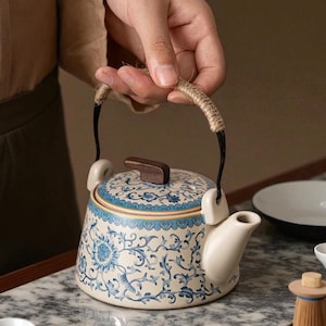 Könnte beinhalten: Keramik-Teekanne mit blau-weißem Blumenmuster. Sie hat einen Holzdeckel und einen mit Schnur umwickelten Griff. Der Ausguss ist weiß, und das Design ist traditionell.