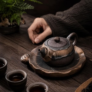 May include: A ceramic tea set comprising a small teapot with a lid, a leaf-shaped tray, and several small teacups. The teapot and tray have a brown and blue glaze. The tea set is on a dark wooden surface.