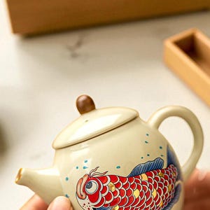 Peut inclure: Petite théière en céramique crème ornée d'un motif de carpe koï. Le poisson est rouge, doré et bleu, avec des points bleus. Elle possède un bouton marron sur le couvercle et une anse incurvée. Idéale pour le thé.