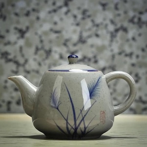 Puede incluir: Una tetera de cerámica con tapa, boquilla y asa. La tetera es de color gris claro con diseños florales azules y un pequeño sello rojo. La tapa tiene un pequeño pomo azul. La tetera está diseñada para preparar té.