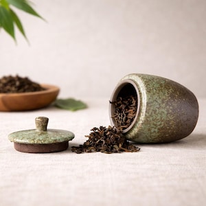 Puede incluir: Una tetera de cerámica verde y marrón con tapa, junto a un montón de hojas de té sueltas. La tetera está volcada, derramando hojas de té. Un cuenco de madera con hojas de té está en el fondo.