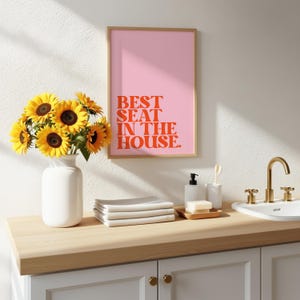 Könnte beinhalten: Badezimmer-Szene mit einem rosa gerahmten Druck mit dem Text "BEST SEAT IN THE HOUSE" in orangefarbenen Buchstaben. Eine weiße Vase mit Sonnenblumen, ein Stapel weißer Handtücher und ein goldener Wasserhahn sind ebenfalls zu sehen.